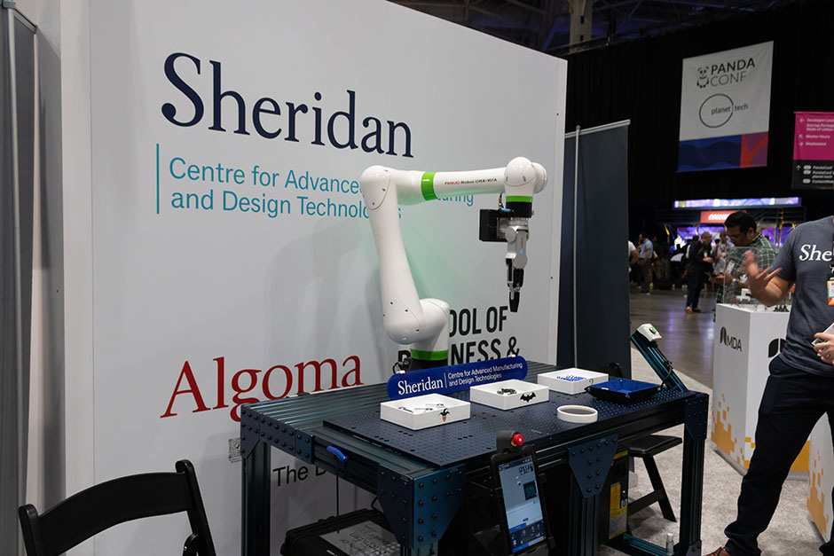A robotic arm on a table perched above three trays with prizes, an engineer gestures at the machine