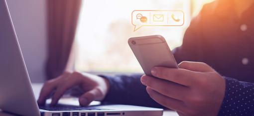 Close-up of someone using a laptop and holding a smartphone, with graphic icons for messaging, email and calling in a bubble above the phone.